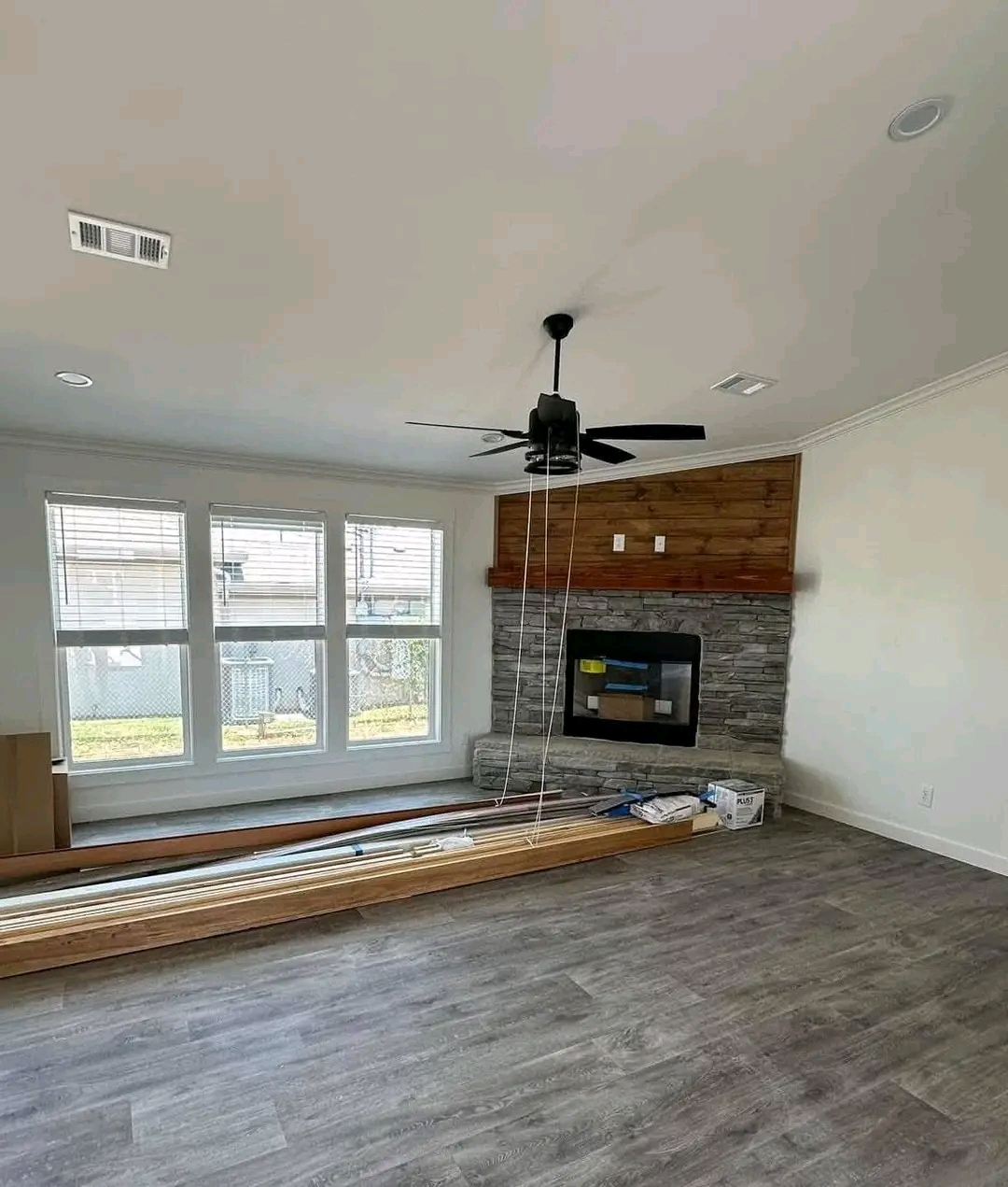 A bright, empty living room with gray flooring, a stone fireplace, and wooden mantel. Large windows let in natural light. Ceiling fan above.