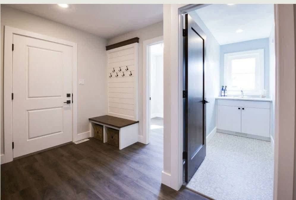 Bright entryway with dark wood floors, a white door on the left, and a bench with hooks. Open door leads to a light blue bathroom with a window.