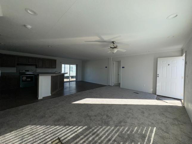 Spacious living room with gray carpet, white walls, and a ceiling fan. Sunlight filters in through a window, casting soft shadows. Kitchen area visible on the left.