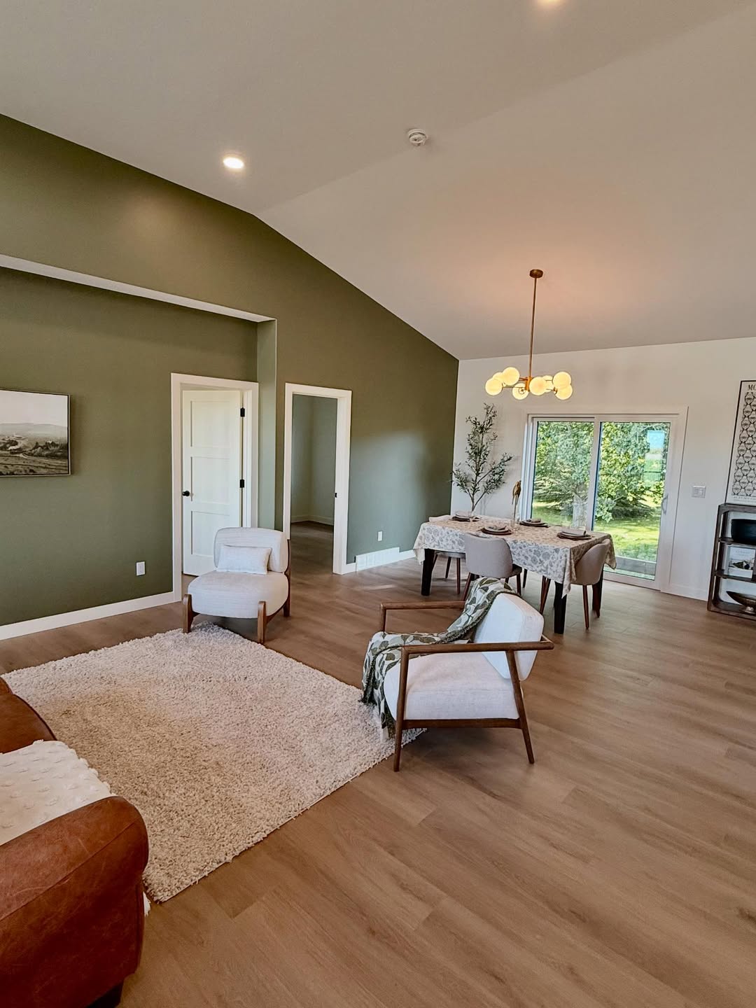 Spacious living and dining area with olive green accent wall, cozy furniture, light wood flooring, and large window overlooking a lush garden.