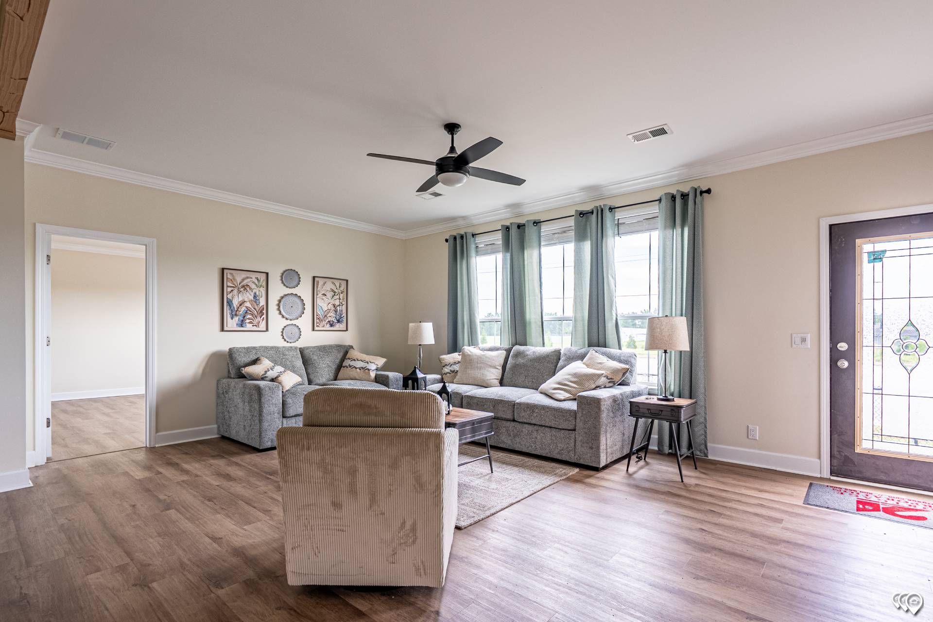 Spacious living room with hardwood floors, grey sofas, and green curtains. Soft lighting creates a cozy atmosphere. Wall art and large window present.