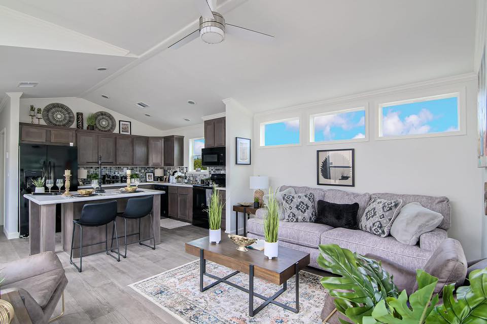 Bright, modern living space with open kitchen. Light gray sofa, patterned cushions, wooden coffee table, and indoor plants. Skylight windows and ceiling fan.