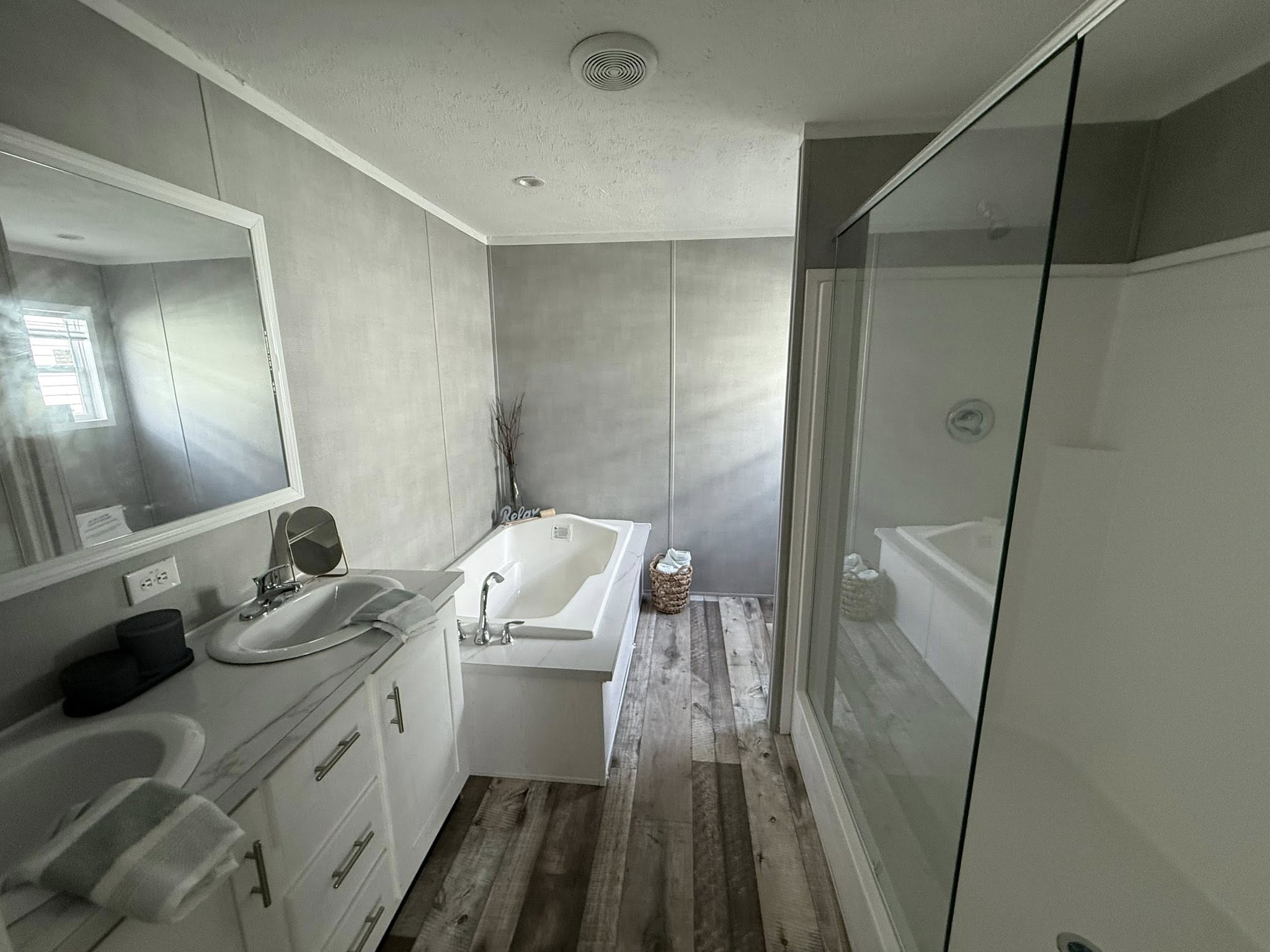 Modern bathroom with wooden floors, featuring a large mirror, double sinks on a white vanity, and a bathtub with a relaxing ambiance.