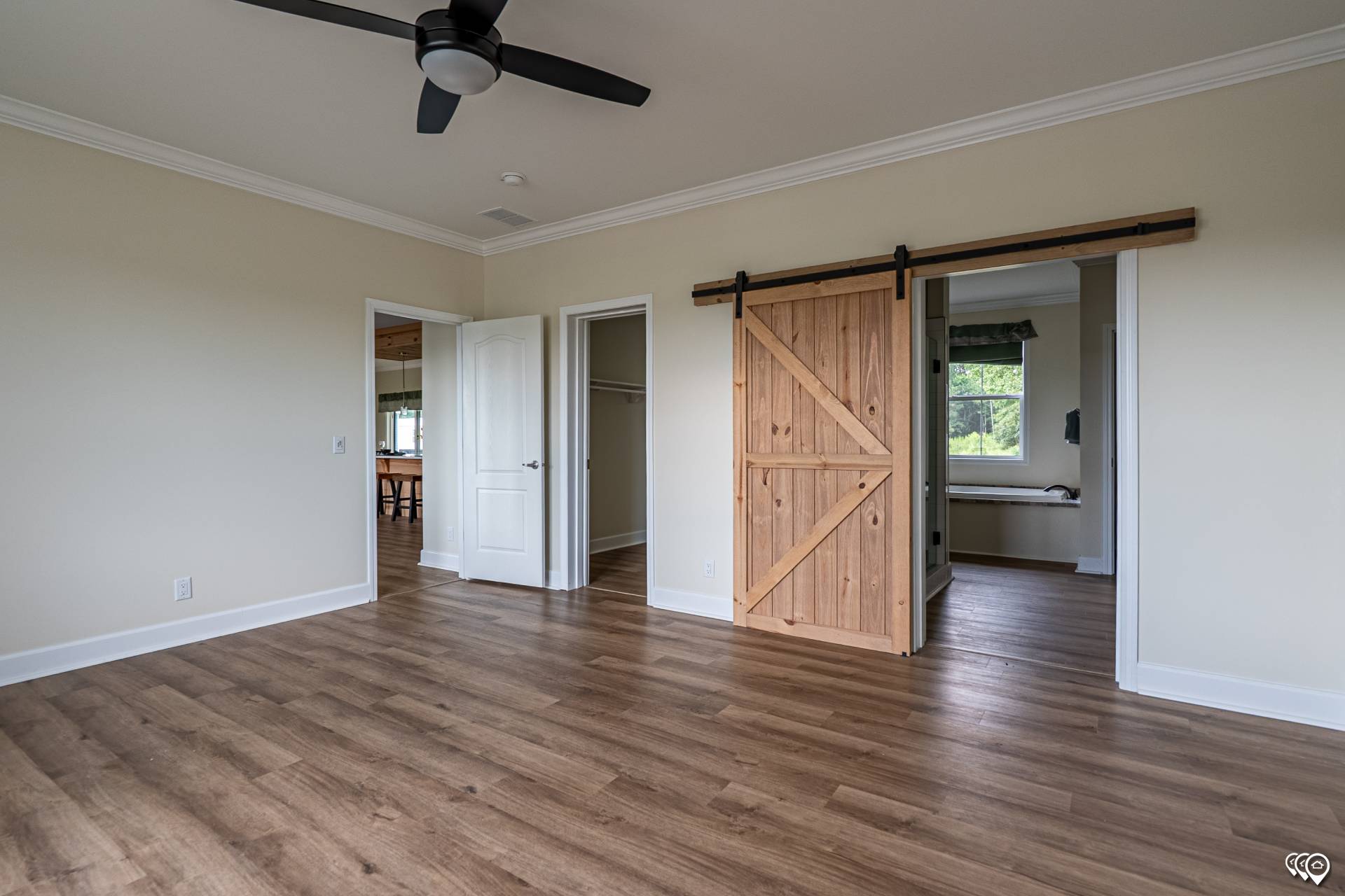 A spacious, empty room with light brown wood flooring and beige walls. It features a ceiling fan, a white door, and a rustic wooden barn door.