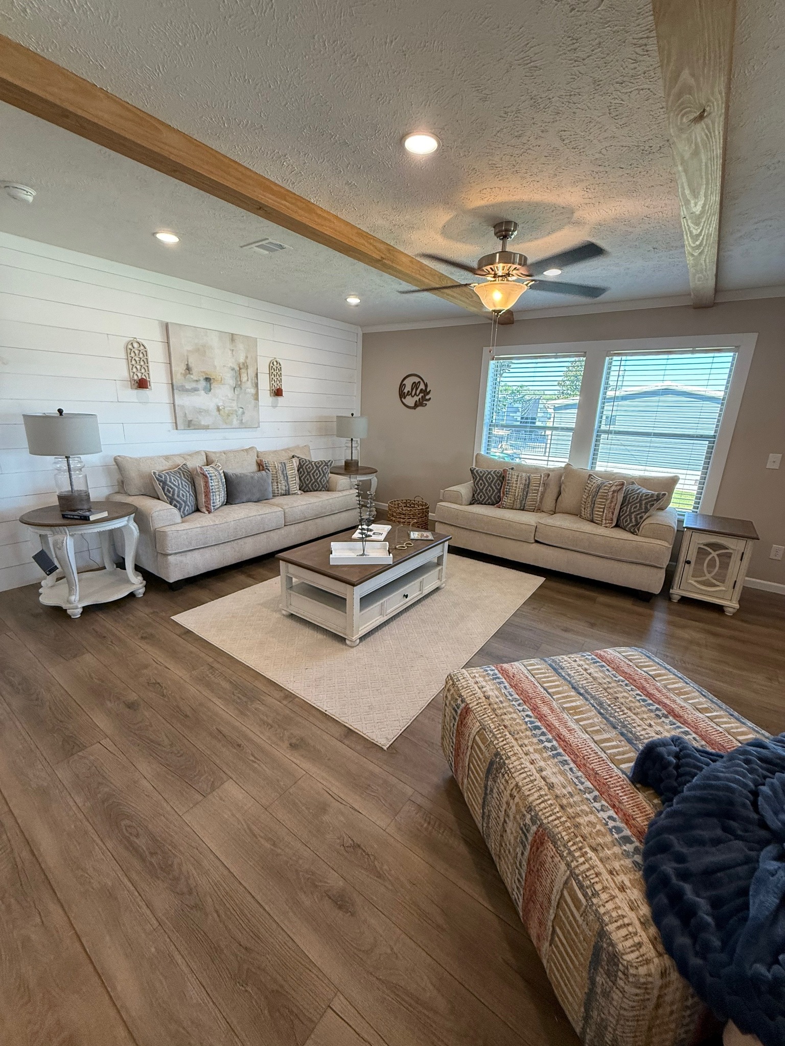Cozy living room with two beige sofas, decorative pillows, wooden coffee table, and lamps. Large windows, wood accents, and warm lighting create a welcoming ambiance.