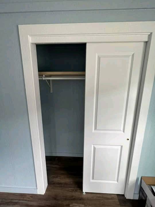 A small closet with a sliding white door, partially open, reveals an empty interior with a single shelf and hanging rod. The room has light blue walls and dark wood flooring, creating a calm, minimalist ambiance.