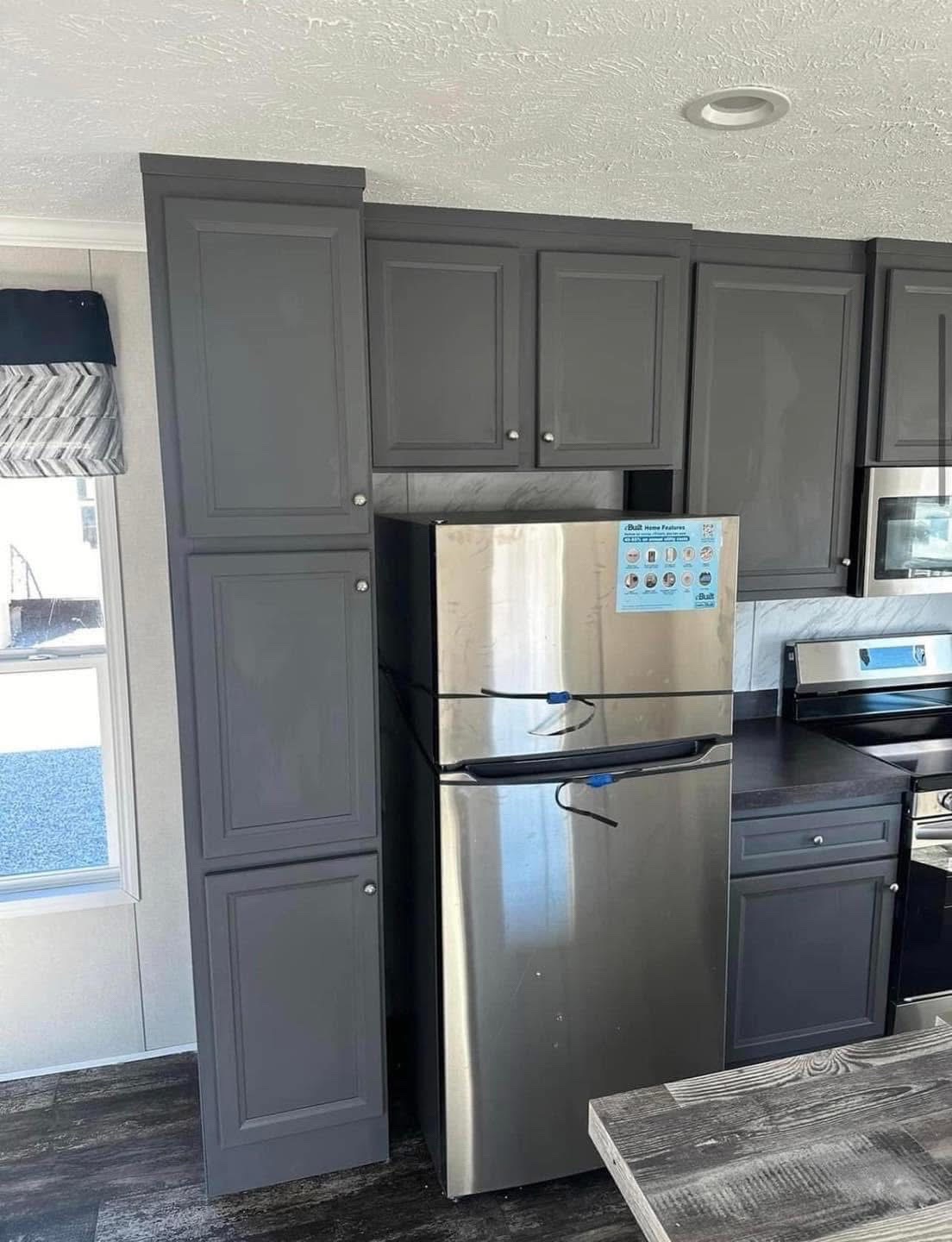 Modern kitchen with dark gray cabinets and a stainless steel refrigerator. Natural light from the window creates a tidy, contemporary feel.