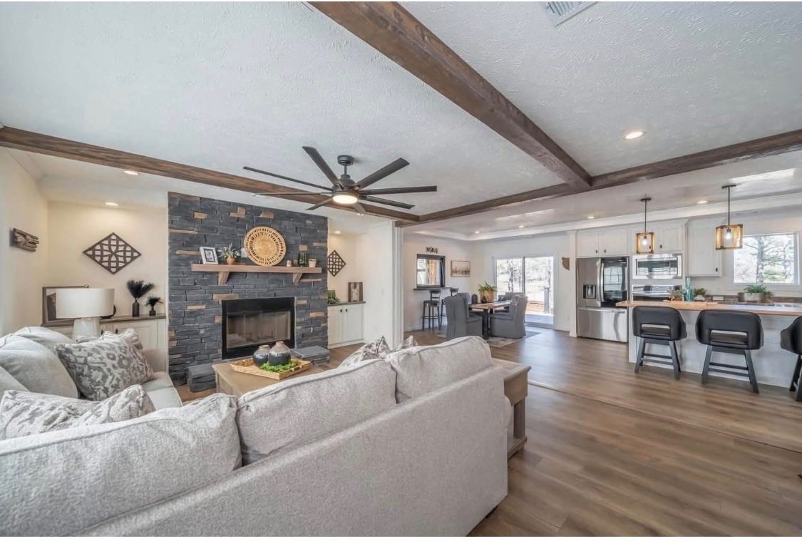 Spacious living room with gray sofas, a stone fireplace, and wooden ceiling beams. Open-plan layout extends to a modern kitchen with a breakfast bar. Cozy and inviting.