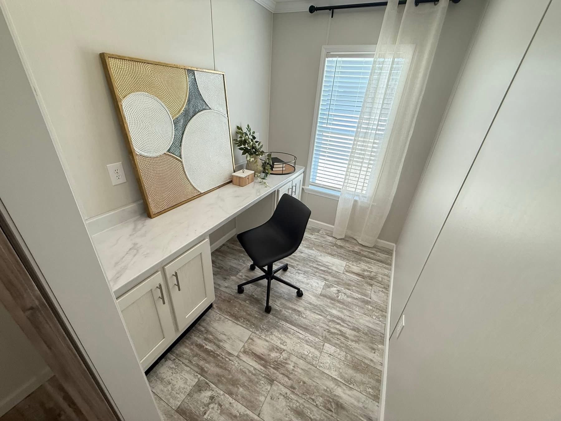 A minimalist home office with a marble desk, black chair, abstract wall art, and a plant on the desk. Sunlight filters through sheer curtains.