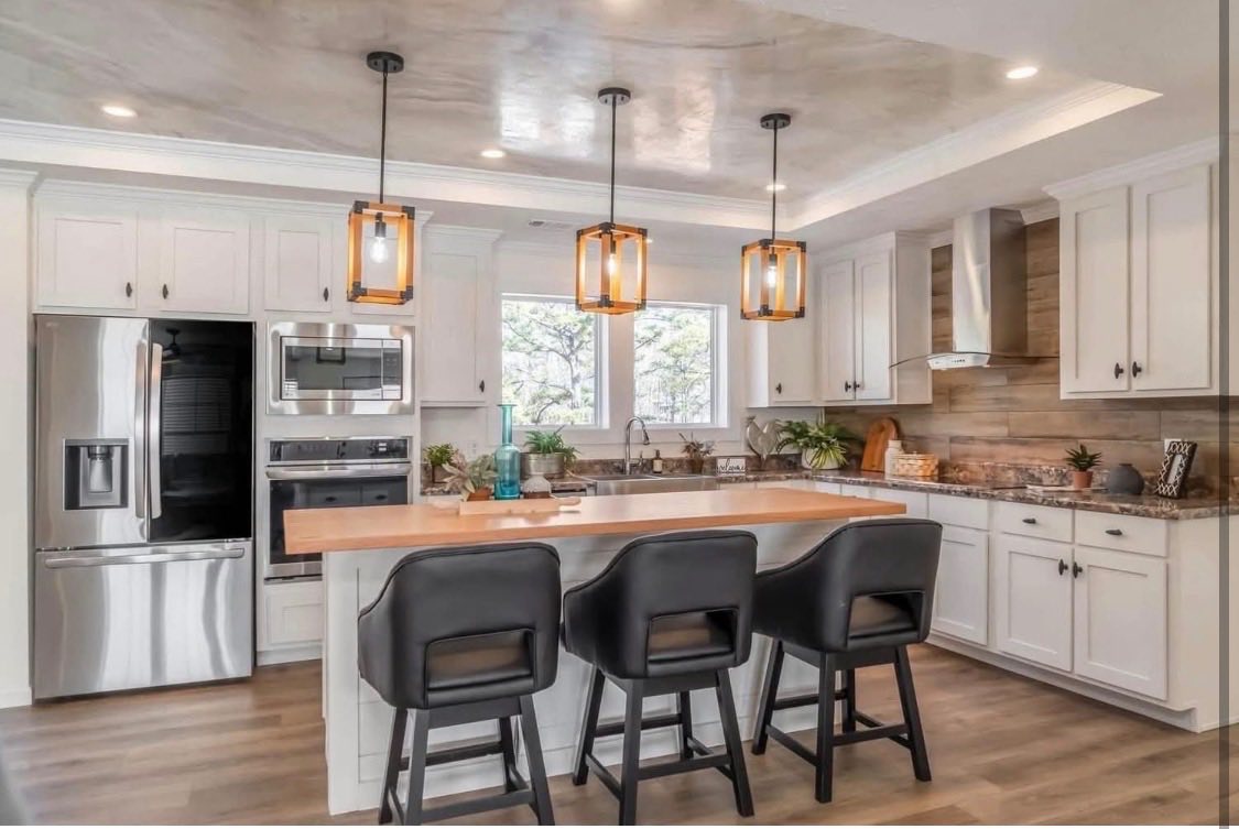 Modern kitchen with white cabinets, stainless steel appliances, and a wooden island. Three pendant lights hang above black chairs, creating a cozy, inviting atmosphere.