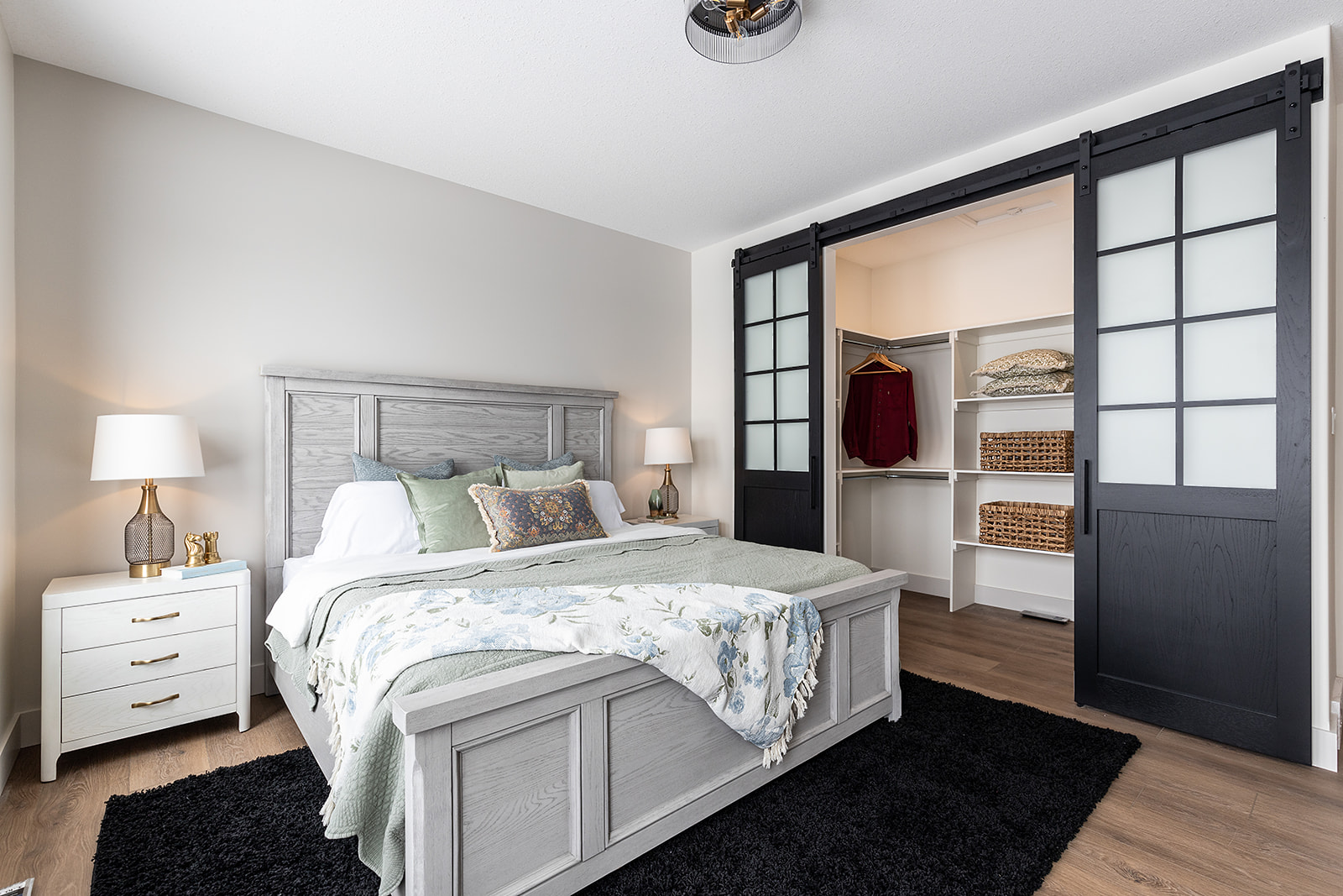 A cozy bedroom features a light wooden bed with floral bedding, flanked by two lamps on white nightstands. A walk-in closet with sliding doors is open, revealing shelves with woven baskets and a hanging garment. The room exudes calmness and modern elegance.