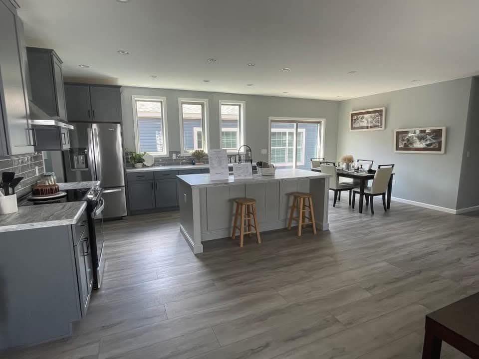 Modern kitchen and dining area with light gray cabinets and flooring. Features an island with stools, stainless steel appliances, and a sunlit dining table. Warm and inviting atmosphere.