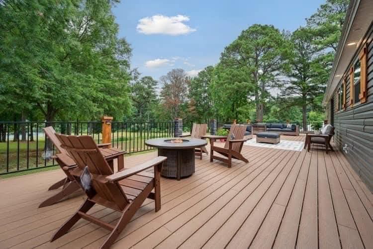 Spacious wooden deck with Adirondack chairs around a fire pit, bordered by a black railing. Trees and a bright sky create a serene atmosphere.