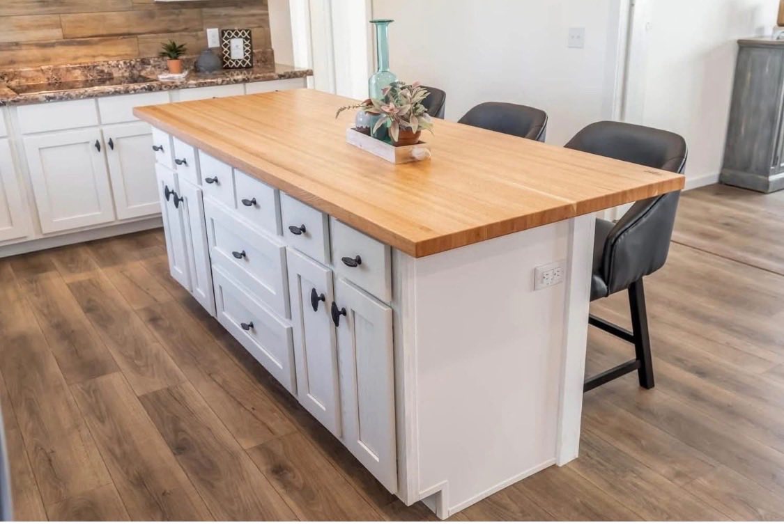 A modern kitchen with a wooden island features white cabinets and black handles. Bar stools line one side, and a vase with plants decorates the top.