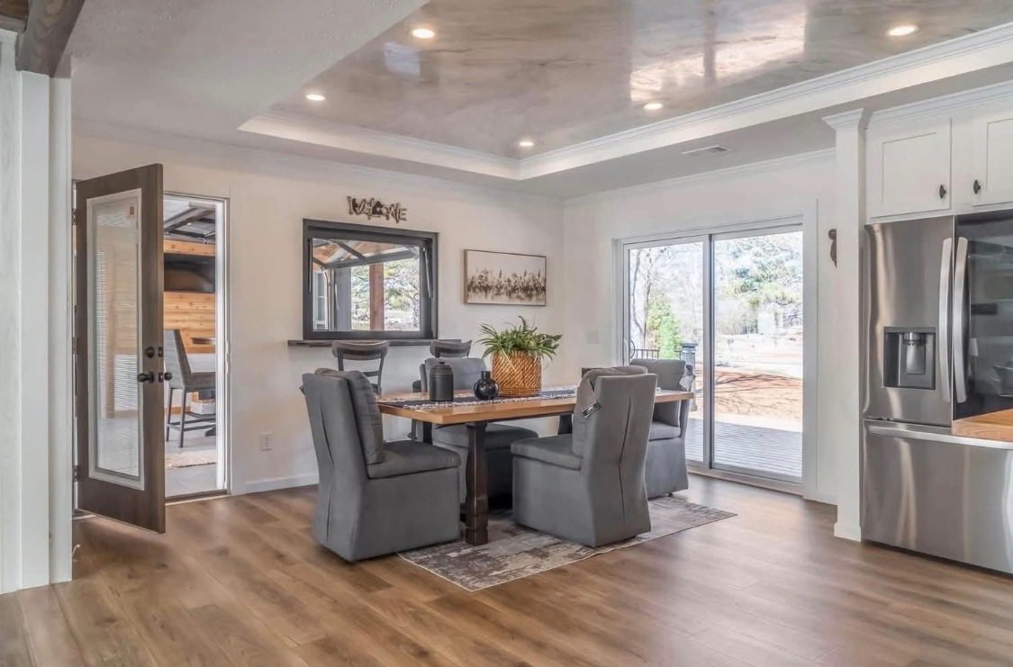 Modern dining room with elegant wood floors, a rectangular table, gray upholstered chairs, and a potted plant centerpiece. Glass doors let in natural light.