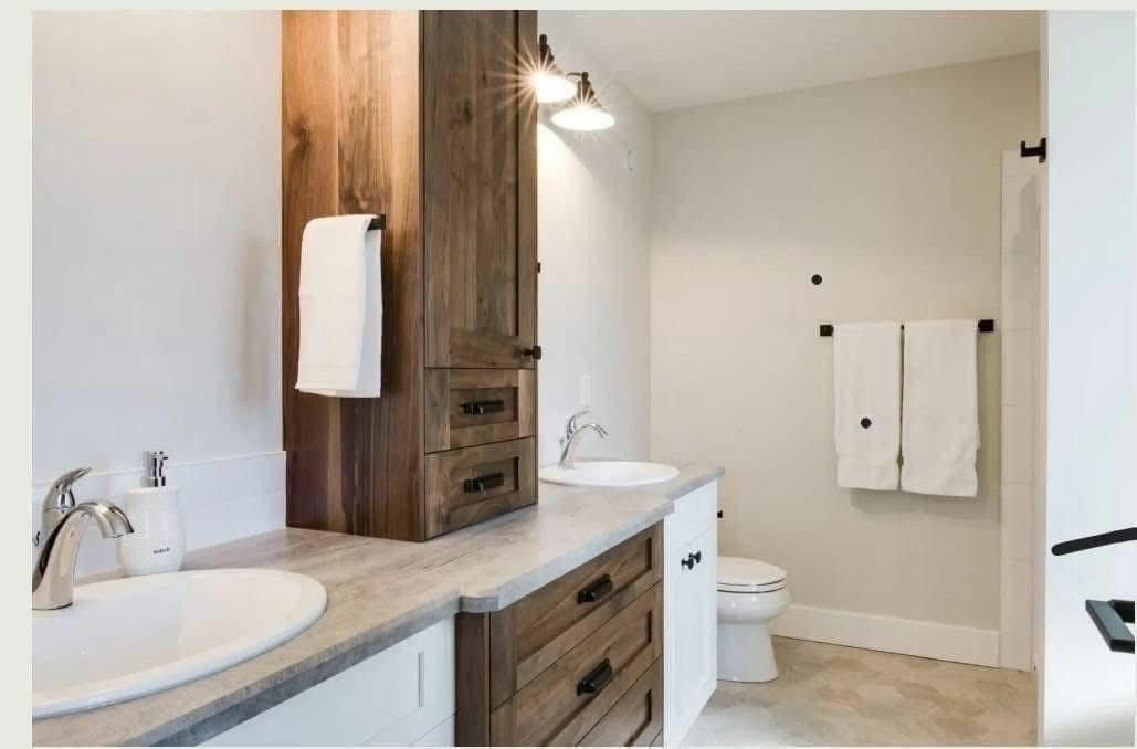 Modern bathroom with dual sinks, each with chrome faucets, and a wooden cabinet. Towel hangs on the wall; neutral tones create a serene feel.