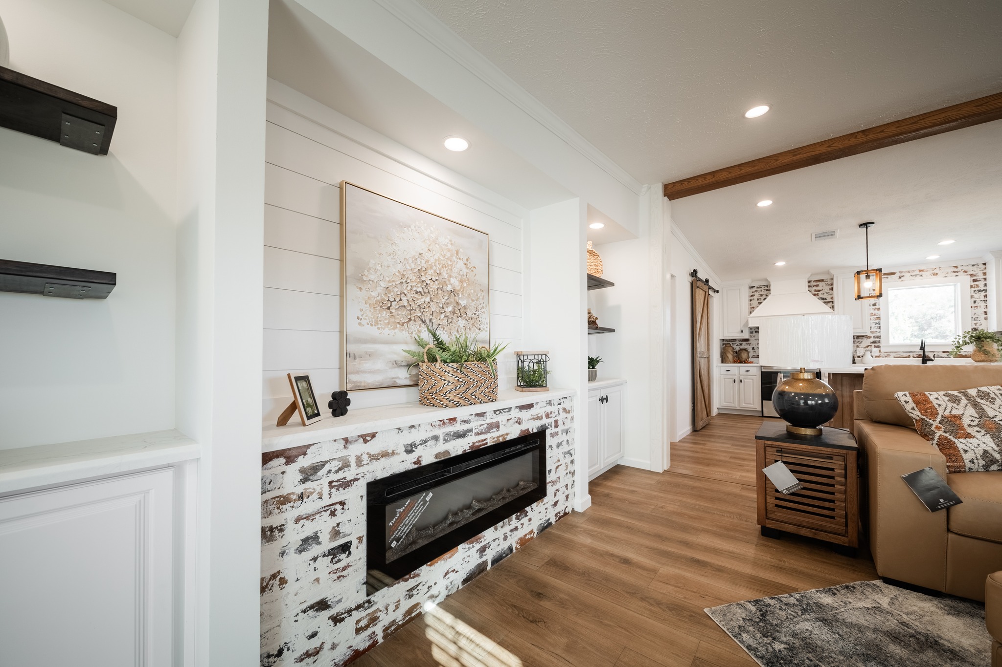 Bright living room with white walls and shiplap, featuring a rustic brick fireplace. A cozy beige couch, wooden floor, and decor create a welcoming feel.