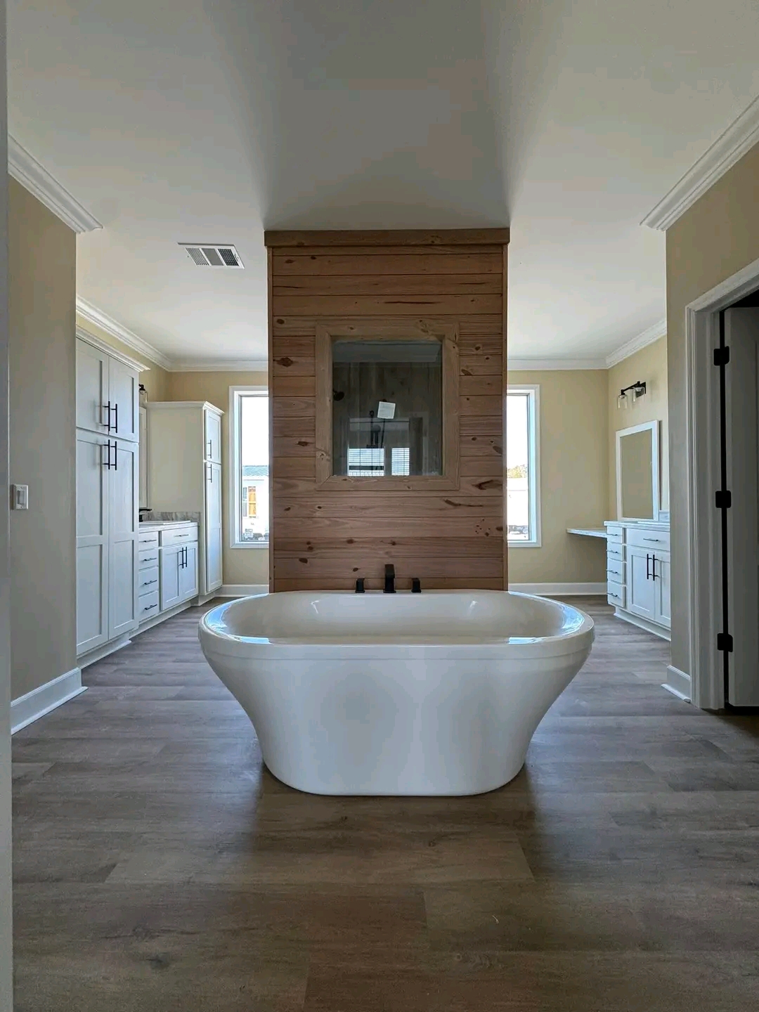 Modern bathroom featuring a central, white freestanding bathtub on wooden flooring. Natural wood paneling, dual vanities, and large windows create an airy, serene space.