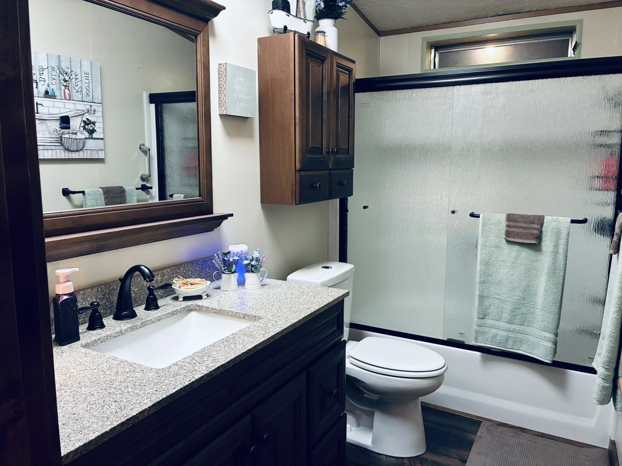 A tidy bathroom with a granite countertop, wooden cabinets, and a large mirror. A glass shower, white toilet, and soft lighting create a cozy atmosphere.