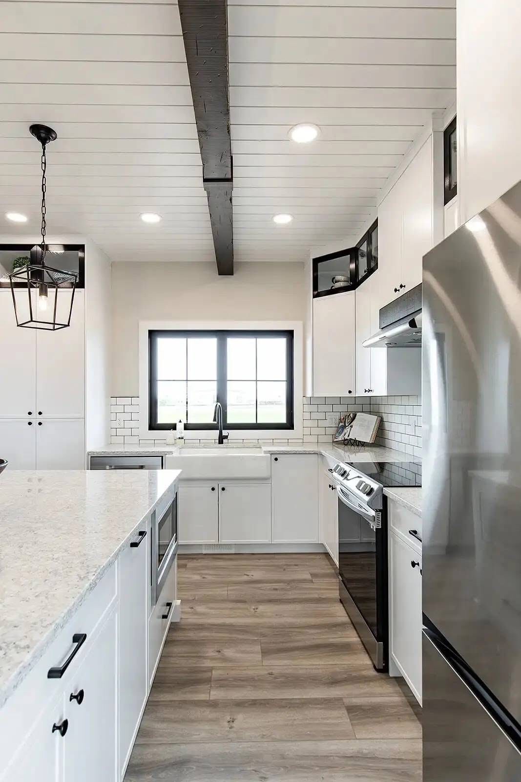 Sleek modern kitchen with white cabinets, subway tile backsplash, and stainless steel appliances. Features a large window, wood floors, and a black beam.