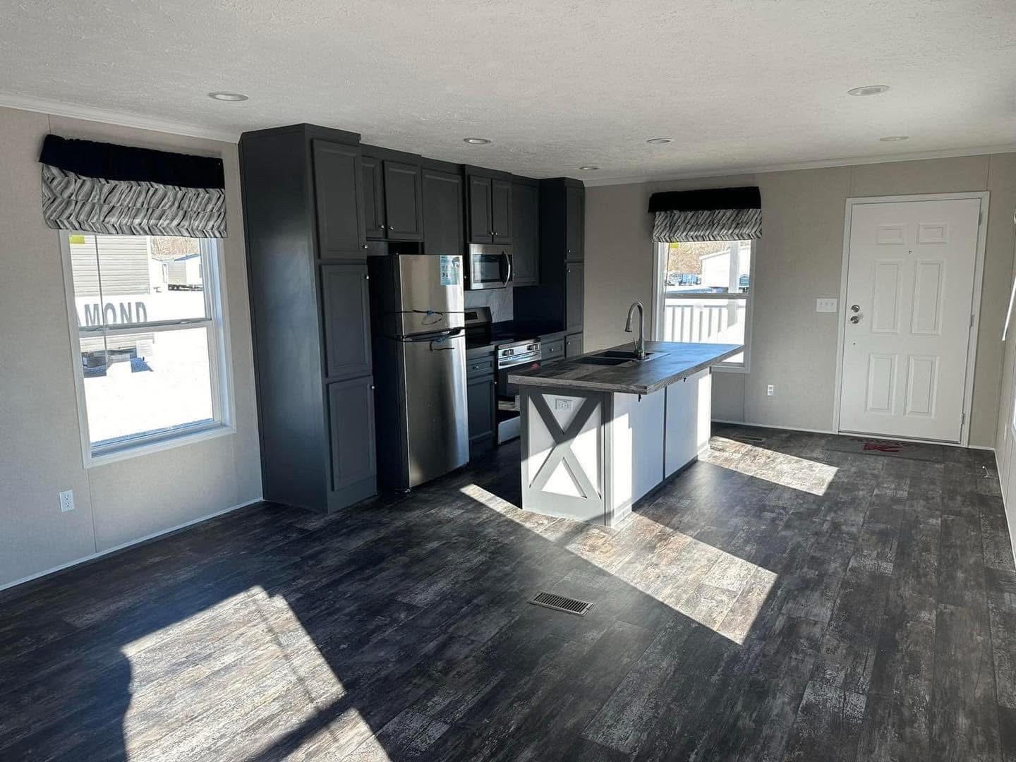 Modern kitchen with dark wood floors and black cabinets, featuring a stainless steel fridge, an island with a sleek sink, and bright natural lighting.