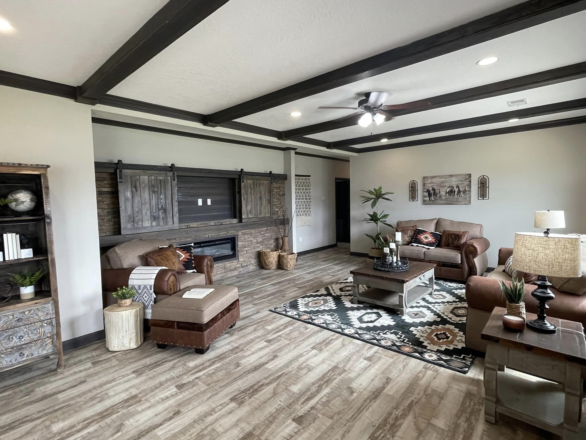 Spacious living room with rustic charm, featuring brown leather sofas, a patterned rug, wooden ceiling beams, and a cozy stone fireplace. Warm, inviting atmosphere.