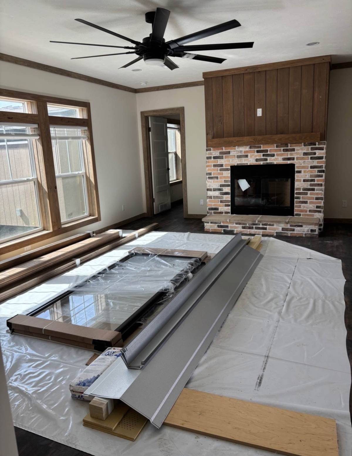 A living room under renovation with a ceiling fan, large windows, brick fireplace, and materials on the floor. Bright and organized.