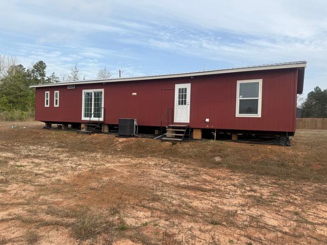 A red mobile home with a white door and windows sits on a dry, grassy area with light brown soil. Trees are visible in the background under a cloudy sky.