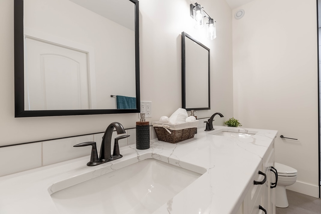 Modern bathroom with dual sinks, black faucets, and marble countertop. Two rectangular mirrors with black frames hang above. A basket with towels and a small plant add decor.