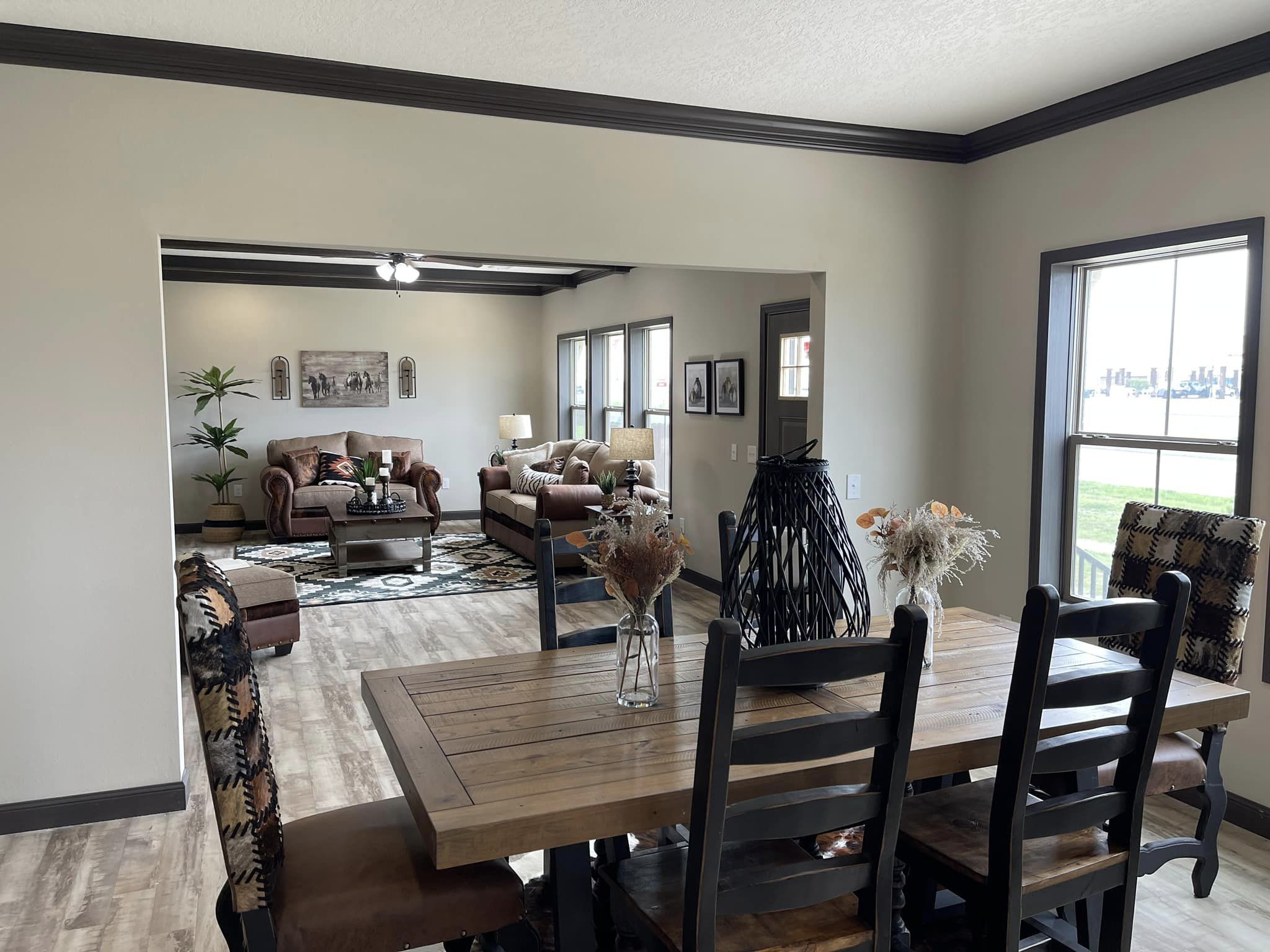 Modern open-concept living and dining area. A wooden dining table with decorative vases is in the foreground. The cozy living room has sofas, plants, and warm lighting.