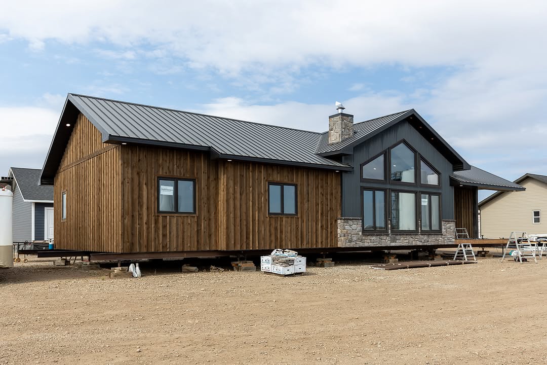 Modern modular home with wood siding and metal roof on a construction site. Large windows and stone chimney suggest a rustic, inviting atmosphere.