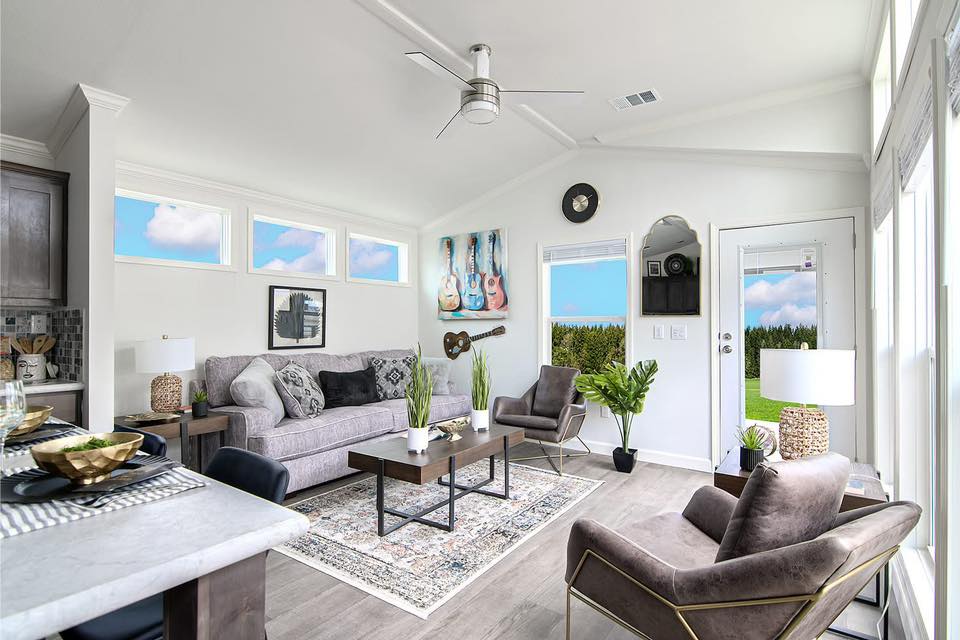 Modern living room with light gray walls, featuring a plush gray sofa, two stylish armchairs, and a coffee table. Large windows with blue skies. Guitar decor and plants add a lively touch.