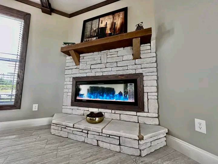 Stone fireplace with a modern electric insert displaying blue flames, set in a cozy living room corner. Wooden mantel holds artwork and decor.