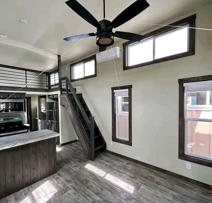 Modern tiny home interior with high ceilings, large windows, and a sleek black ceiling fan. Features a loft with stairs, kitchenette, and wood flooring.