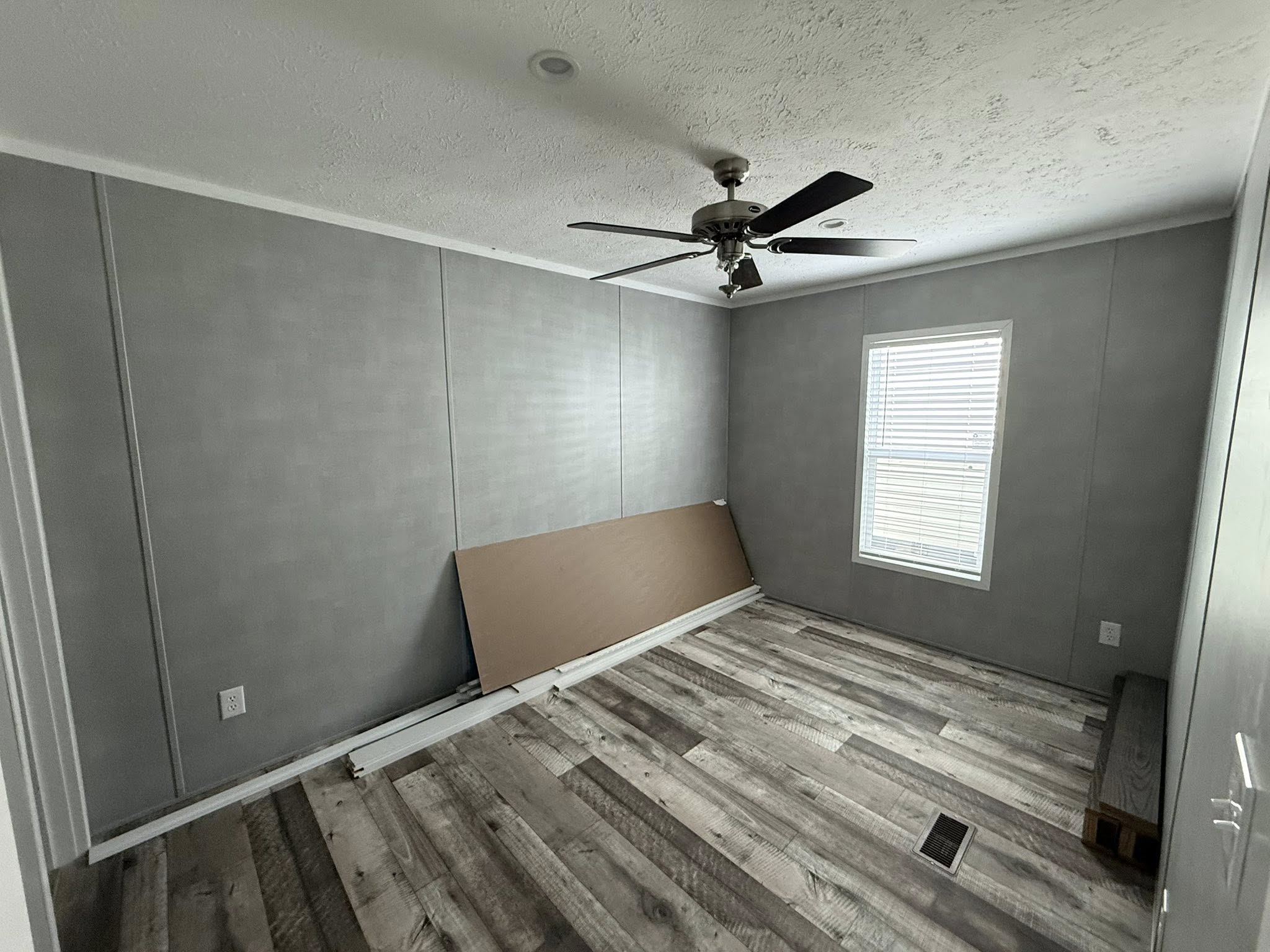 A small, empty room features gray walls, a ceiling fan, and a single window with blinds. A wooden board leans against the wall, and the floor has gray wood-like planks. The room feels calm and minimal.