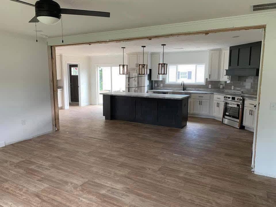 Open floor plan with a modern kitchen; dark island, pendant lights, and wooden floor. White cabinets, black hood, bright window, and ceiling fan.