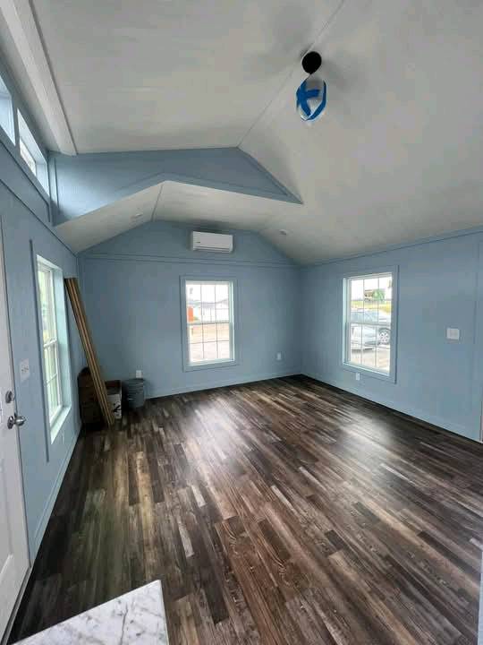 Bright, empty room with light blue walls and high, slanted ceiling. Wooden floor, two windows, and an overhead light fixture create a spacious, calm atmosphere.