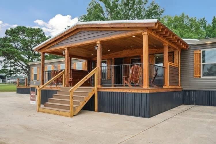 A cozy, rustic wooden porch with steps leads up to a modern mobile home. Surrounded by trees, the porch includes railings and a rocking chair, conveying a warm, inviting atmosphere.