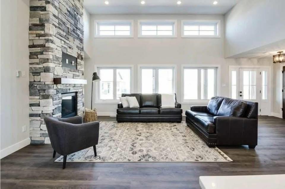 Spacious living room with high ceilings, large windows, and a modern stone fireplace. Features two black leather sofas, a gray armchair, and a patterned rug.