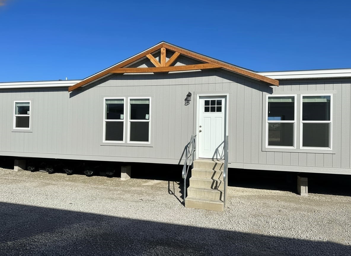 A gray modular home under a clear blue sky, featuring a front door with steps, rustic wooden trim, and several windows, creating a welcoming tone.