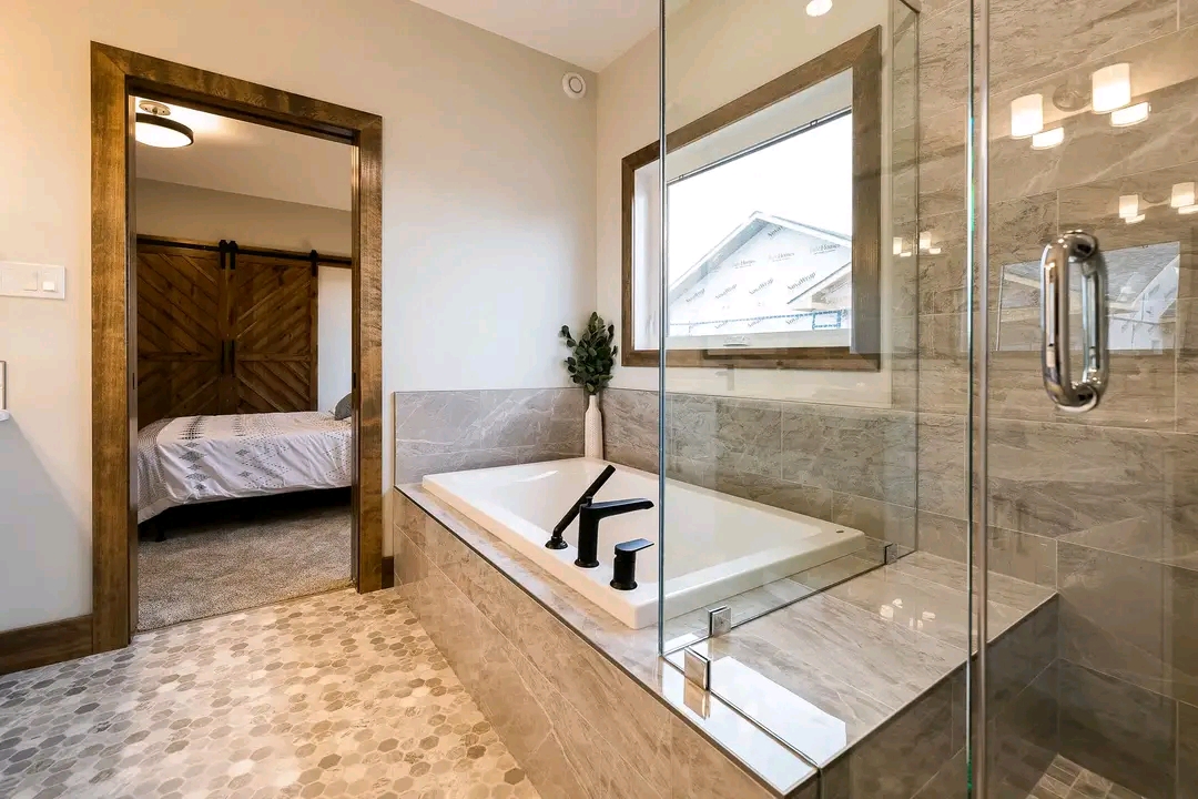 Modern bathroom featuring a glass-enclosed shower, sleek bathtub with black fixtures, stone-tile floor, and a view into a bedroom with a barn door.