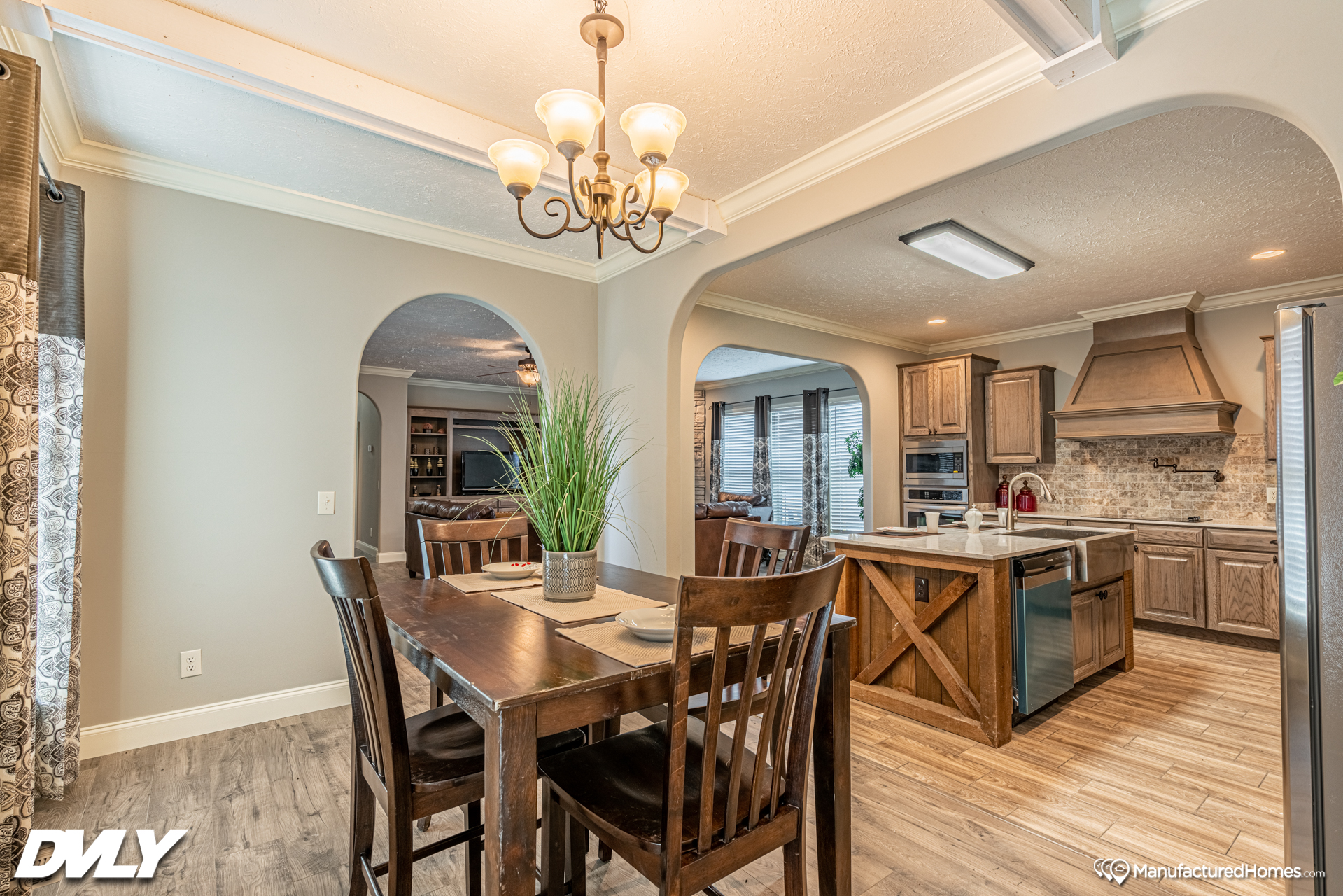 Bright dining area with a dark wood table set, under a classic chandelier. Open archways lead to a rustic kitchen with wood cabinetry and an island.
