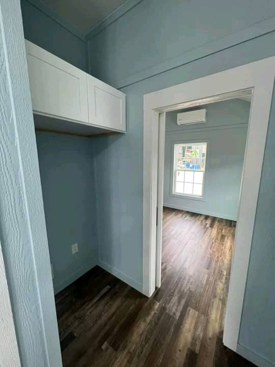 Compact blue room with white cabinets and wood flooring leads to a bright, larger room with a window, suggesting an airy, fresh atmosphere.