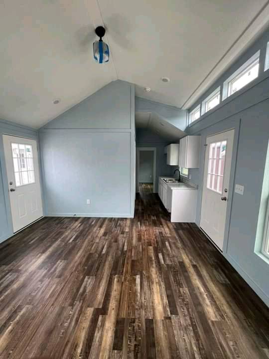 A minimalist room with a vaulted ceiling, light blue walls, and dark wood flooring, featuring two doors, windows, and a small kitchenette.