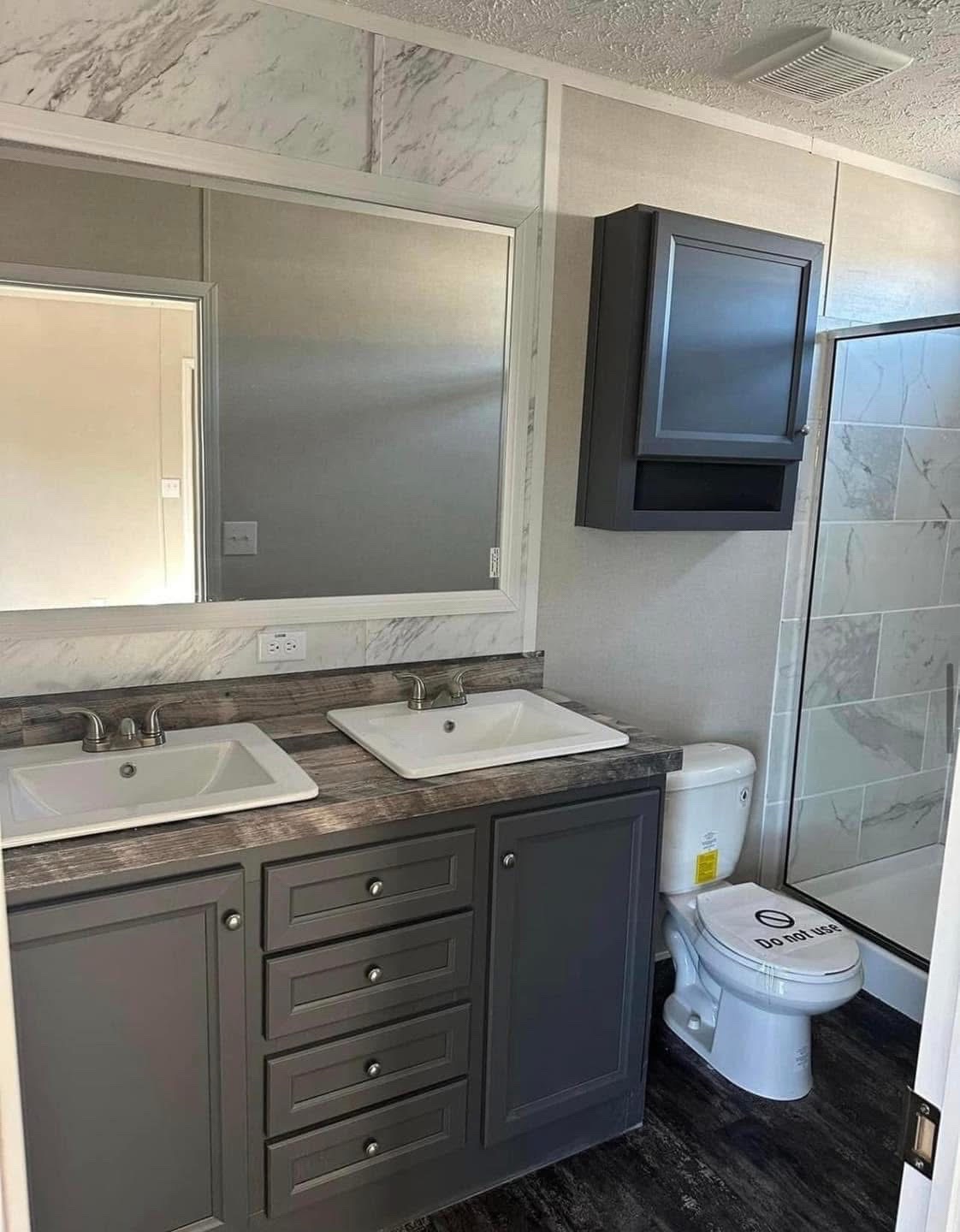 Modern bathroom with dual sinks, large mirror, dark wood cabinets, and a toilet labeled "Do Not Use." A shower with marble tiles is visible.