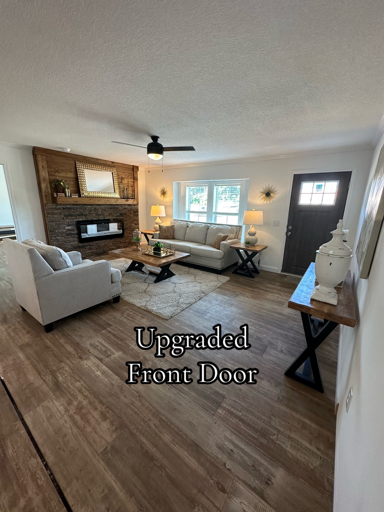 Spacious living room with a cozy, modern vibe. Features a stone-accented fireplace, light sofas, wooden coffee table, and large windows by an upgraded front door.
