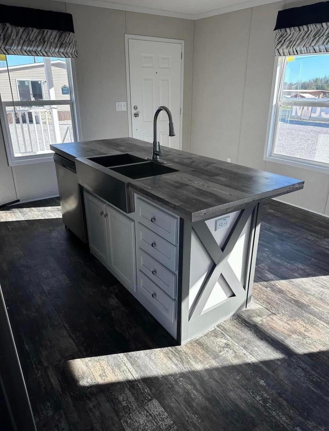 A modern kitchen with a center island featuring a dark countertop, built-in sink, and cabinetry. Sunlight streams through large windows, creating a bright, airy atmosphere.