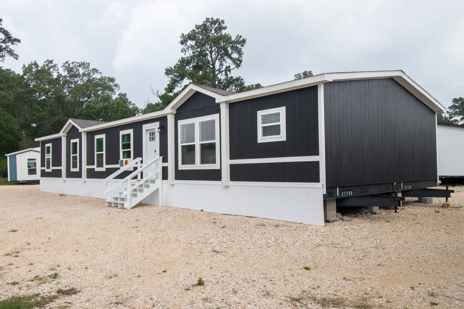 A modern, black and white mobile home with multiple windows sits on a gravel lot. Trees are visible in the background under a cloudy sky.