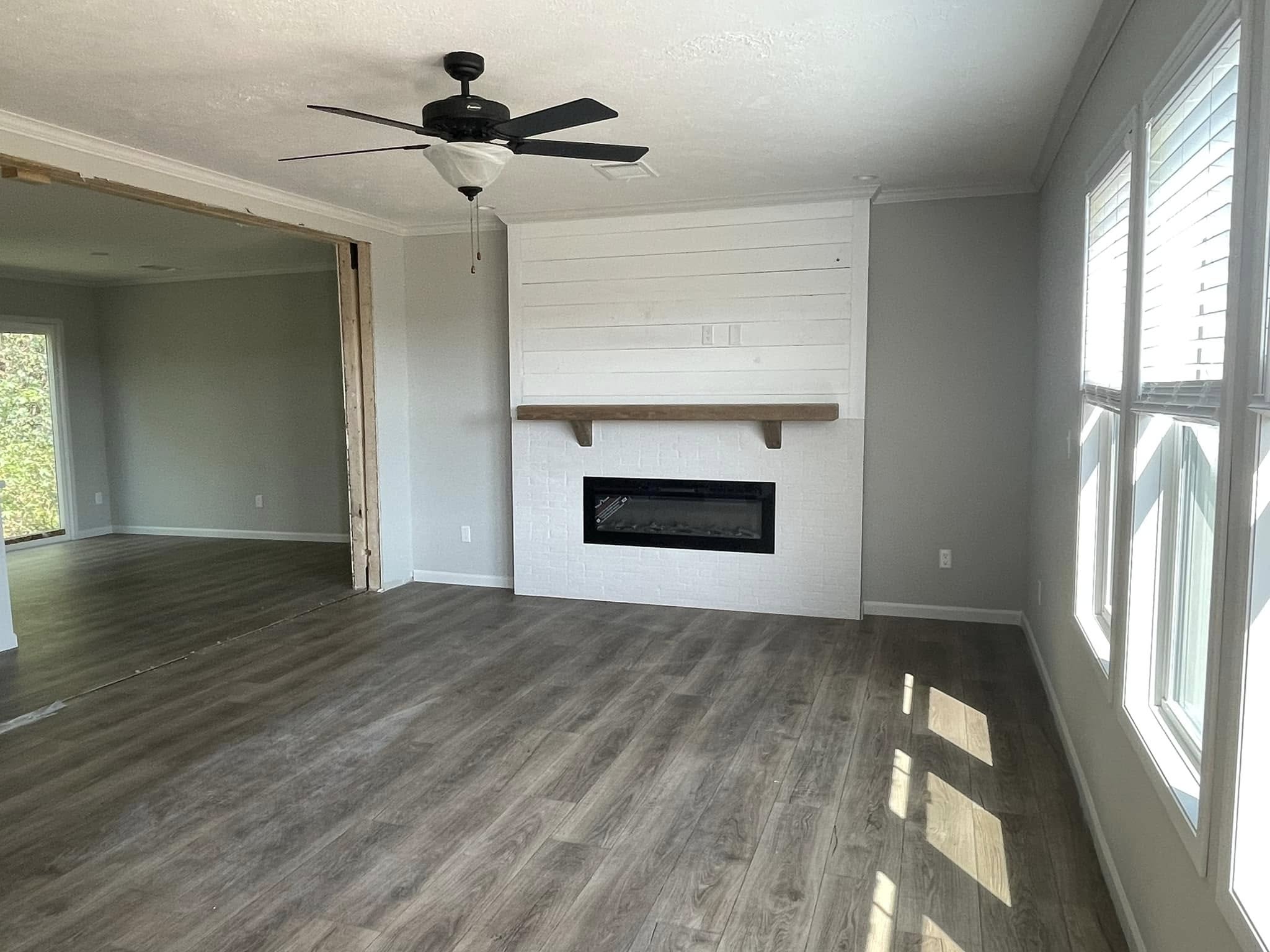 Spacious living room with gray walls, large windows, and wood flooring. Features a white shiplap accent wall with a modern fireplace and a ceiling fan.