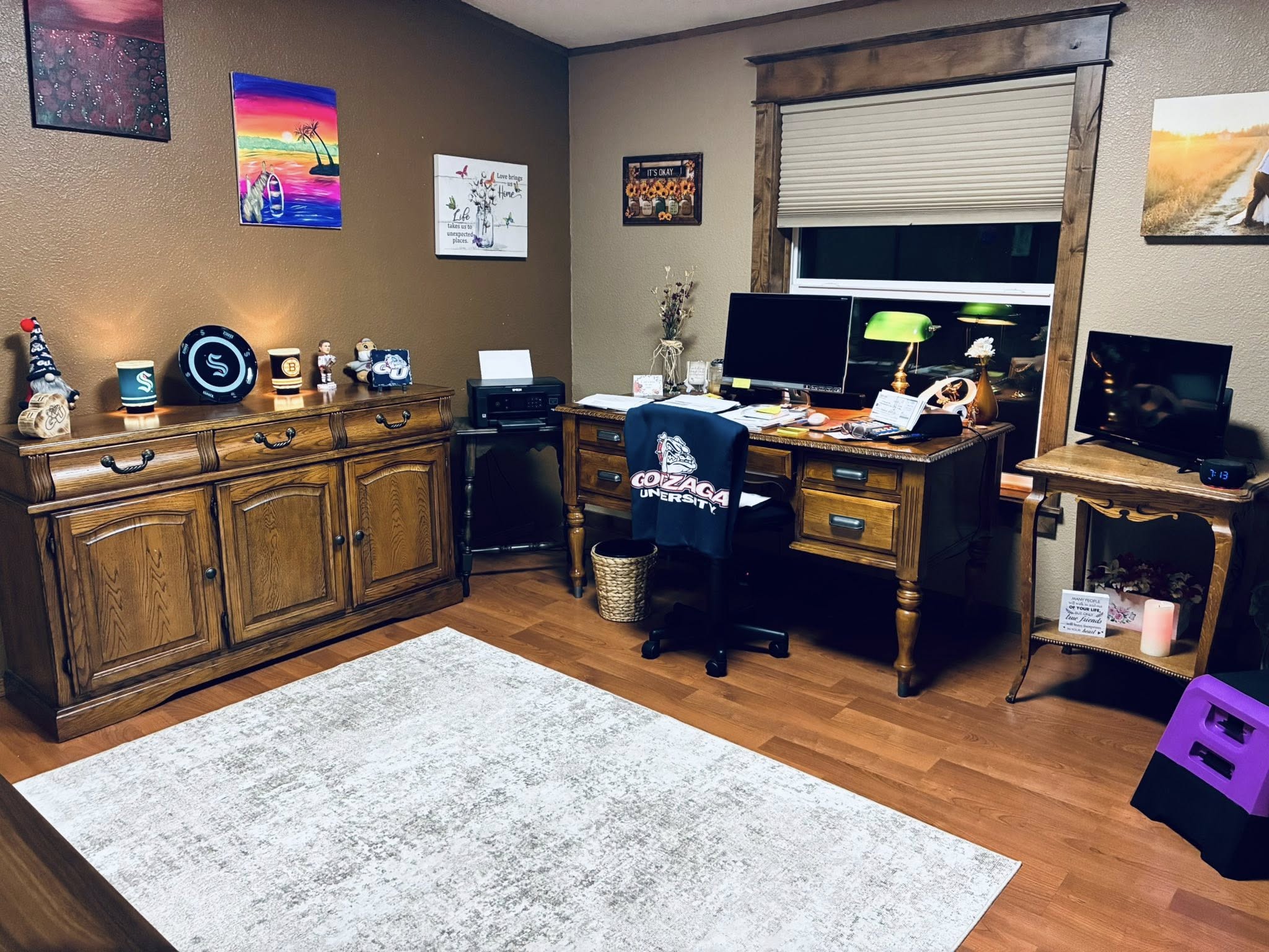 A cozy home office features a wooden desk and cabinets, adorned with lamps and decorations. Art and certificates hang on warm-toned walls, creating an inviting atmosphere.